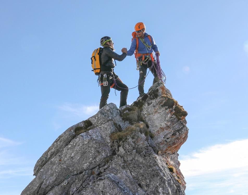 Two individuals standing on a mountain peak, overlooking a vast landscape of hills and valleys under a clear blue sky.